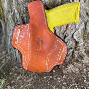 May include: A brown leather holster with white stitching and a yellow gun inside. The holster has a small American flag patch on the front.