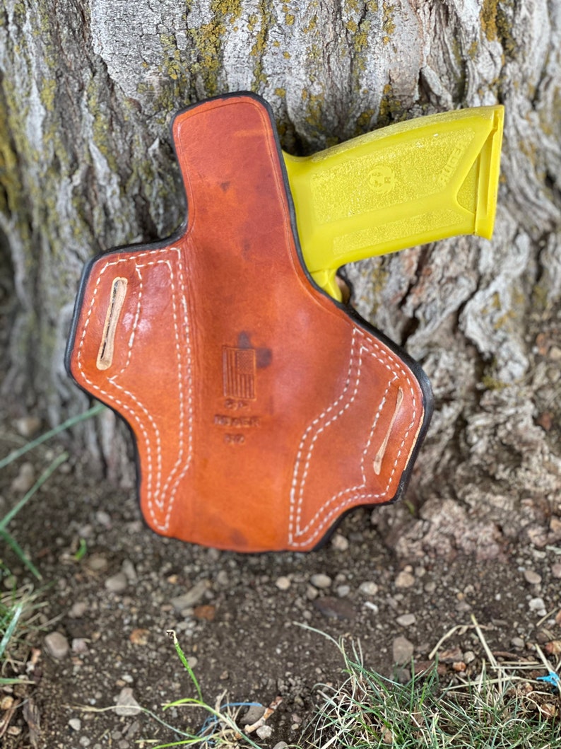 May include: A brown leather holster with white stitching and a yellow gun inside. The holster has a small American flag patch on the front.