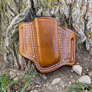 Może przedstawiać: Brązowa skórzana kabura z białymi szwami. Kabura jest przeznaczona do przechowywania małego przedmiotu, takiego jak nóż lub latarka.