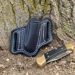 May include: A black leather knife sheath with white stitching, designed to hold a folding knife. The sheath is lying on the ground next to a black-handled folding knife with a brass bolster.