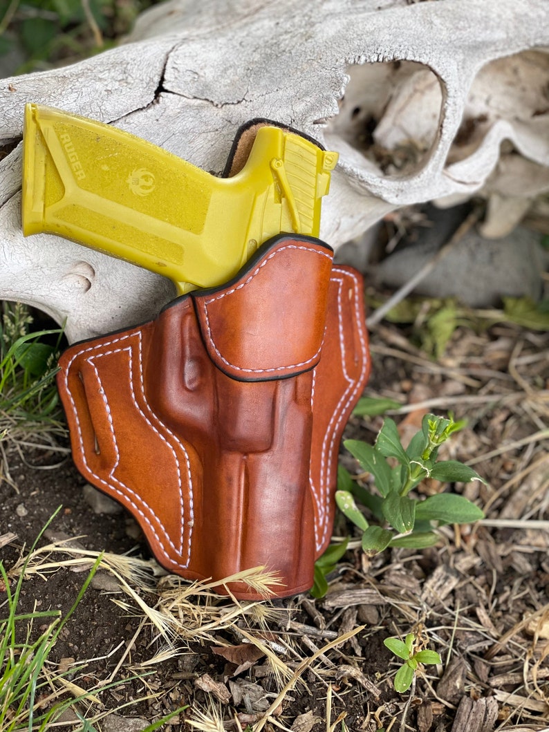 May include: A brown leather holster with white stitching, designed to hold a yellow handgun with the word "RUGER" printed on the side.