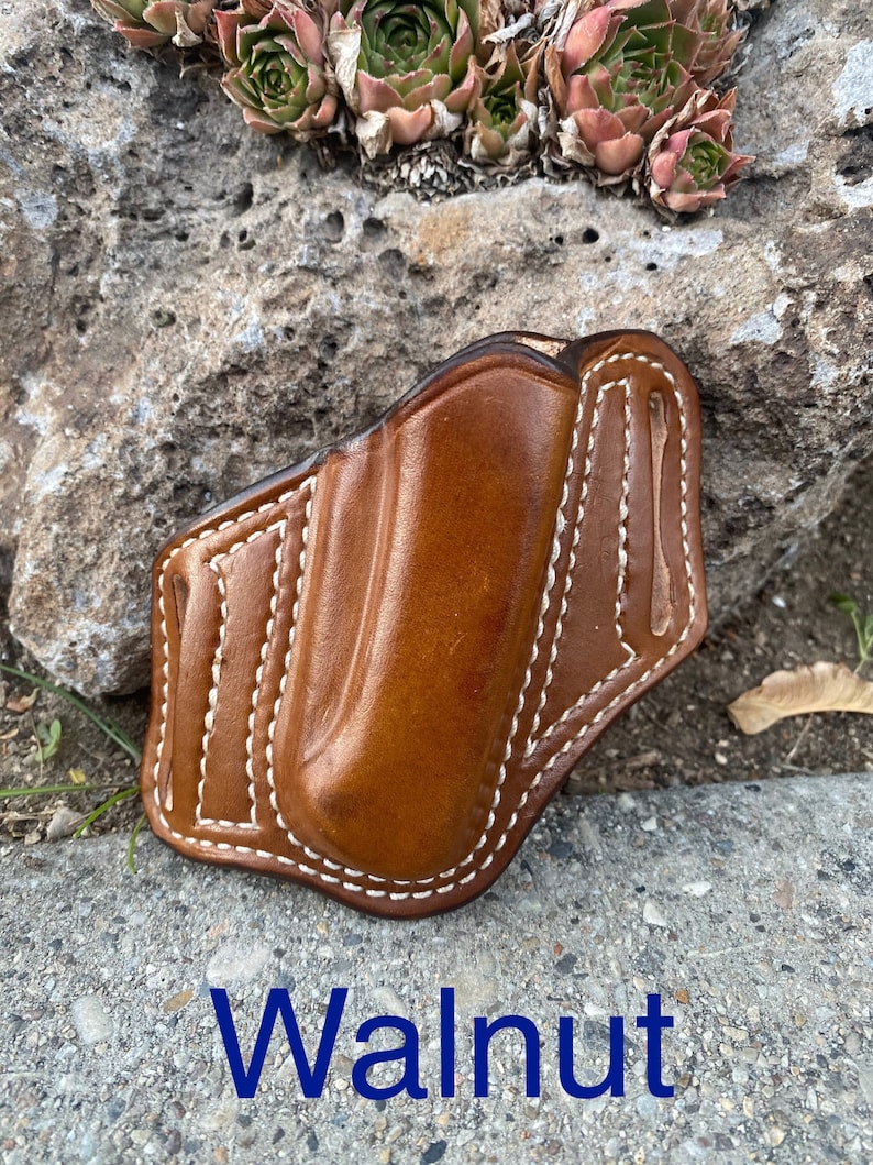 May include: A brown leather holster with white stitching. The holster is designed to hold a small pocket knife or other small tool. The text "Walnut" is printed below the holster.