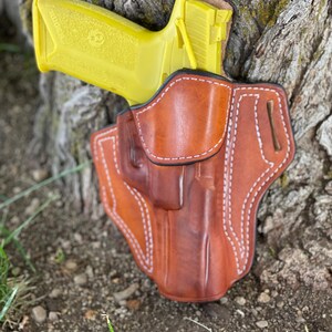 May include: A yellow handgun in a brown leather holster. The holster has white stitching and a belt loop.