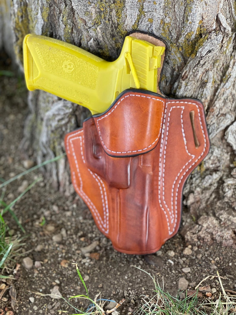 May include: A yellow handgun in a brown leather holster. The holster has white stitching and a belt loop.