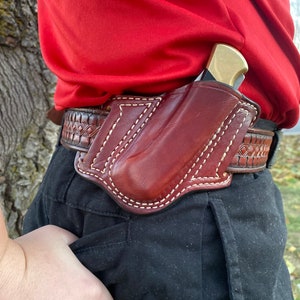 May include: A brown leather holster with a stitched design, worn on a person's belt. The holster is designed to hold a small knife or other tool.