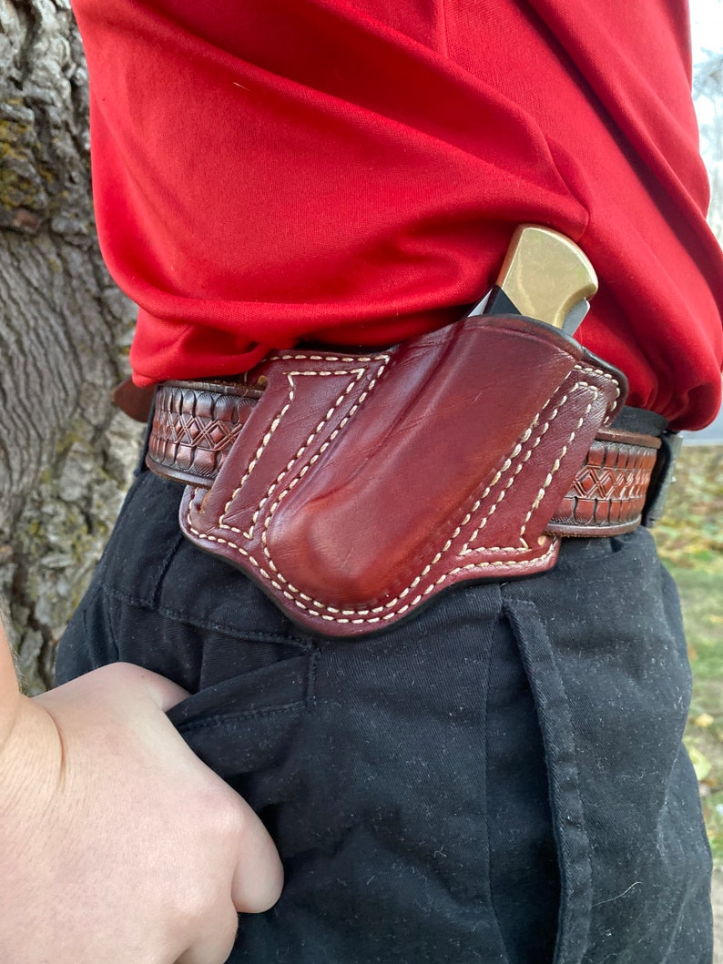 May include: A brown leather holster with a stitched design, worn on a person's belt. The holster is designed to hold a small knife or other tool.