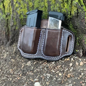 May include: A brown leather magazine holder with two black magazine pouches. The holder has white stitching and is sitting on a bed of small rocks.