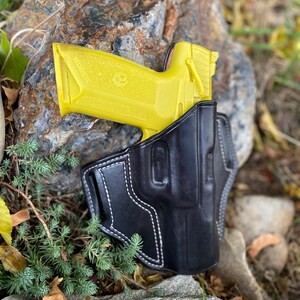May include: A yellow handgun in a black leather holster with white stitching. The holster is on a rock with green foliage.
