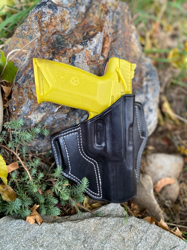 May include: A yellow handgun in a black leather holster with white stitching. The holster is on a rock with green foliage.