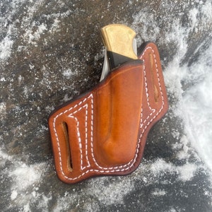 May include: A brown leather knife sheath with white stitching. The sheath is designed to hold a folding knife with a brass handle.