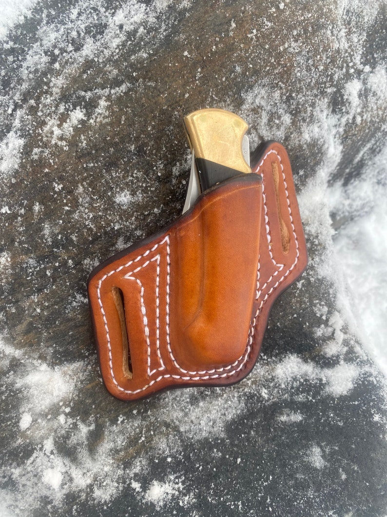 May include: A brown leather knife sheath with white stitching. The sheath is designed to hold a folding knife with a brass handle.
