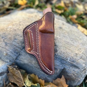 May include: A brown leather knife sheath with white stitching. The sheath is sitting on a rock with leaves in the background.
