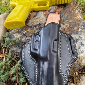 May include: A black leather holster with white stitching for a handgun. The holster is lying on a rock with a yellow plastic handgun next to it.