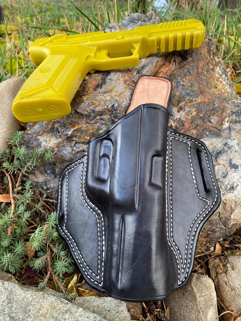 May include: A black leather holster with white stitching for a handgun. The holster is lying on a rock with a yellow plastic handgun next to it.