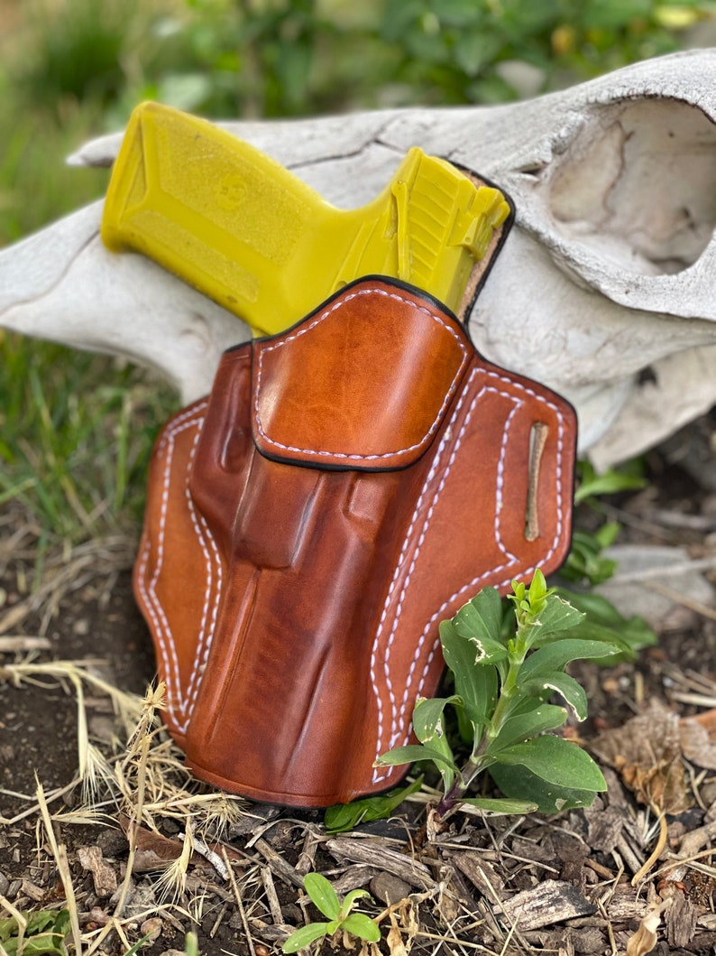 May include: A brown leather holster with white stitching, designed to hold a yellow handgun. The holster is resting on a white animal skull and brown dirt.