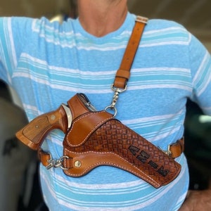 Pode incluir: Um coldre de ombro de couro marrom com um padrão tecido e o nome "Chris" gravado no lado. Um revólver prateado está no coldre.