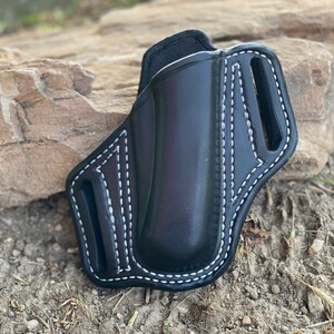 May include: A black leather holster with white stitching. The holster has a belt loop and a flap that covers the opening.