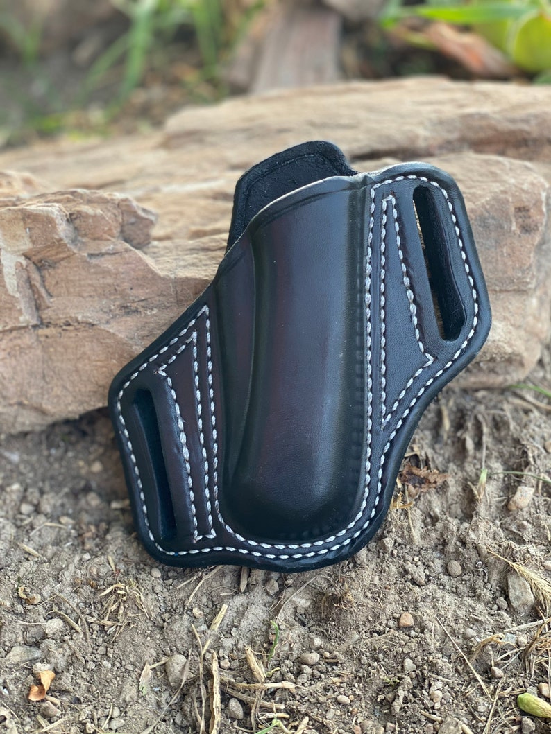 May include: A black leather holster with white stitching. The holster has a belt loop and a flap that covers the opening.