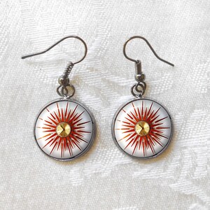 May include: A pair of round earrings with a white background and a red sunburst design. The center of each earring features a gold-colored accent. The earrings have silver-colored hooks.
