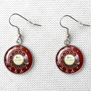 May include: A pair of retro-style earrings featuring a rotary phone dial design. The earrings have a dark red background with white numbers and letters, and the center dial has the text "AREA CODE 613 503-0571". The earrings are suspended from silver-colored hooks.