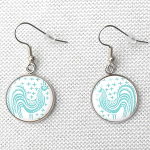 May include: A pair of silver earrings with a round, white background and a turquoise blue rooster design.