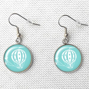 May include: A pair of silver-toned dangle earrings. Each earring features a round, light blue pendant with a white hot air balloon design. The balloon has a detailed pattern and is suspended with white lines. The earrings have a simple hook closure.