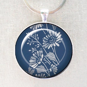 May include: A round pendant necklace with a silver-tone metal frame. The pendant features a dark blue background with a white floral design. The necklace is displayed on a light beige fabric.
