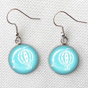 May include: A pair of round earrings with a light blue background and a white hot air balloon design. The earrings have silver-colored hooks.