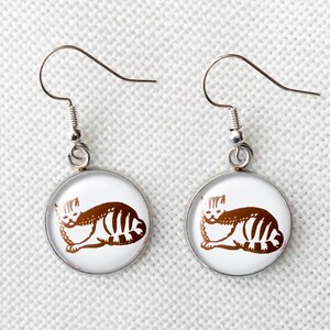 May include: A pair of silver-toned cat earrings. Each earring features a round, white background with a brown cat illustration. The cat is lying down with striped markings. The earrings have a hook closure.