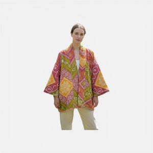 May include: A colorful, patterned kimono-style jacket with a geometric design in shades of pink, yellow, and green. The jacket has wide sleeves and a relaxed fit. The model is wearing a white top and light yellow pants.