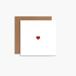 May include: A white greeting card with a small, red glitter heart in the center, paired with a brown envelope. The card is square-shaped, and the heart adds a touch of sparkle to the minimalist design. Perfect for expressing love or affection.