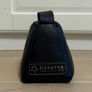 May include: A black leather bag with a handle. The bag has a rectangular shape with a trapezoidal base. The brand name "DEPSTER" is displayed in gold lettering on a rectangular patch, along with the phrase "celebrate your style."