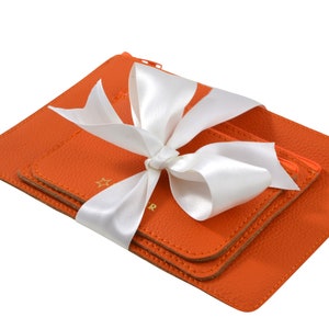 Op de afbeelding: Drie oranje leren etui's met een wit satijnen lint dat in een strik is gebonden. De etui's zijn op elkaar gestapeld en hebben een gouden sterontwerp.