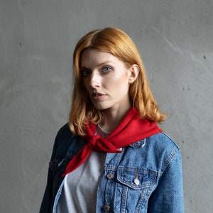 May include: A woman with red hair wearing a denim jacket and a red scarf. She is standing in front of a gray wall.