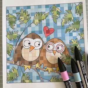 May include: A coloring page featuring two brown owls with large eyes sitting on a branch, surrounded by green leaves and a blue checkered background, with a red heart above them. Three Winsor & Newton Promarker pens are placed on the right side.