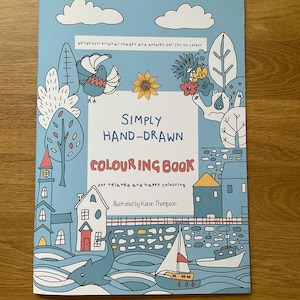 Könnte beinhalten: Ein Malbuch mit hellblauem Einband und dem Text Simply Hand-Drawn Colouring Book. Die Illustration auf dem Cover zeigt eine Küstenszene mit Häusern, einem Segelboot, einem Hai, einem Vogel, einer Sonnenblume und Bäumen im einfachen Linienstil.