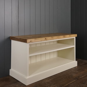 May include: A white wooden cabinet with two shelves and a natural wood top. The cabinet has a rectangular shape and is painted in a creamy white color. The top is made of light-colored wood. The cabinet is designed for storage.