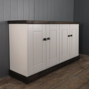 May include: A white cabinet with four doors and dark brown trim. The cabinet has black knobs on each door. The cabinet is sitting on a wooden floor.