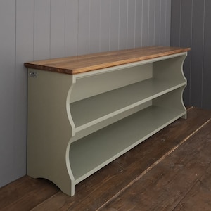 May include: A light green wooden shoe rack with two shelves and a natural wood top. The shoe rack has a curved front and is sitting on a wooden floor.