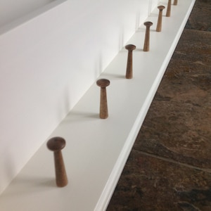 May include: A white wooden shelf with a row of eight wooden hooks. The hooks are stained a dark brown color and are evenly spaced along the shelf.