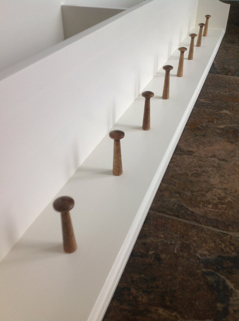 May include: A white wooden shelf with a row of eight wooden hooks. The hooks are stained a dark brown color and are evenly spaced along the shelf.