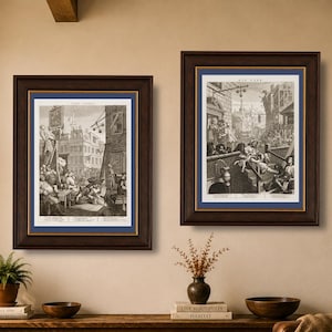 Op de afbeelding: Twee ingelijste zwart-wit prenten met bruine lijsten en blauwe passe-partouts hangen aan een beige muur. De prenten tonen gedetailleerde historische scènes. De kamer heeft een houten console, een leren bank en een grote plant in een pot.