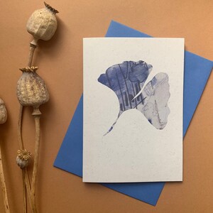 May include: A white card with a blue ginkgo leaf design. The card is on a blue envelope and a brown surface.