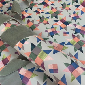 May include: A roll of wrapping paper with a geometric pattern of colourful stars on a light grey background. The paper is rolled up and tied with a grey ribbon.