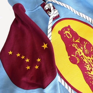 May include: A light blue hoodie with maroon sleeves featuring a constellation of yellow stars. The front of the hoodie has a large yellow circle with a maroon bear silhouette.