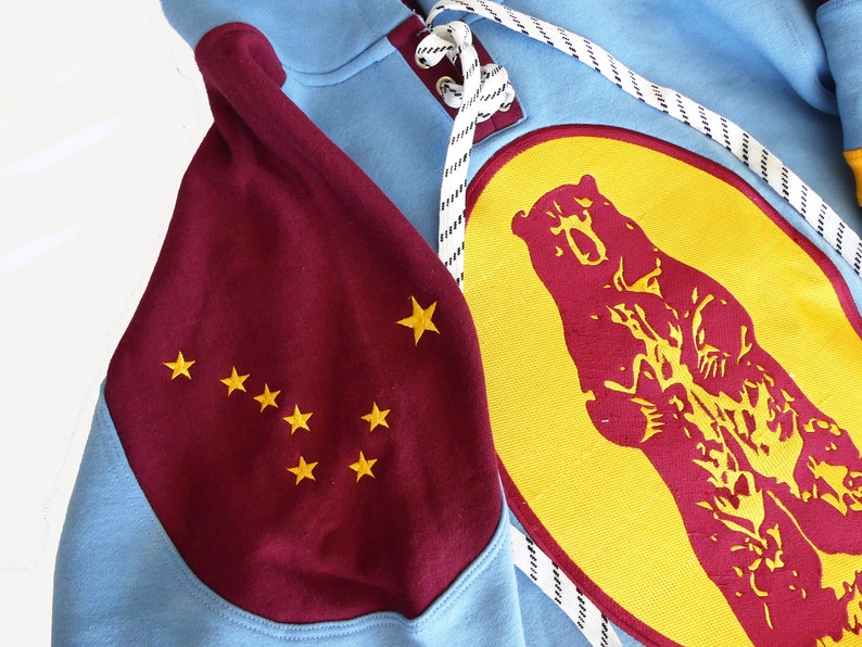 May include: A light blue hoodie with maroon sleeves featuring a constellation of yellow stars. The front of the hoodie has a large yellow circle with a maroon bear silhouette.