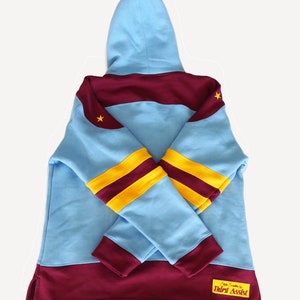 May include: A light blue hooded sweatshirt with maroon and yellow stripes on the sleeves. The sweatshirt has a maroon band across the chest and a maroon band at the bottom. The sweatshirt has a small yellow star on the left shoulder and a small yellow star on the right shoulder. The sweatshirt has a small maroon patch on the bottom right side with the text "Officially Sanctioned by Third Assist".