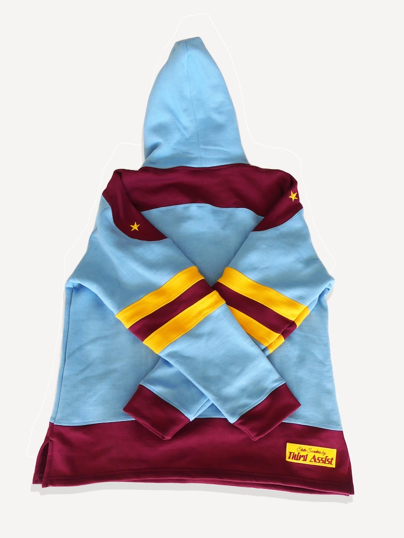 May include: A light blue hooded sweatshirt with maroon and yellow stripes on the sleeves. The sweatshirt has a maroon band across the chest and a maroon band at the bottom. The sweatshirt has a small yellow star on the left shoulder and a small yellow star on the right shoulder. The sweatshirt has a small maroon patch on the bottom right side with the text "Officially Sanctioned by Third Assist".