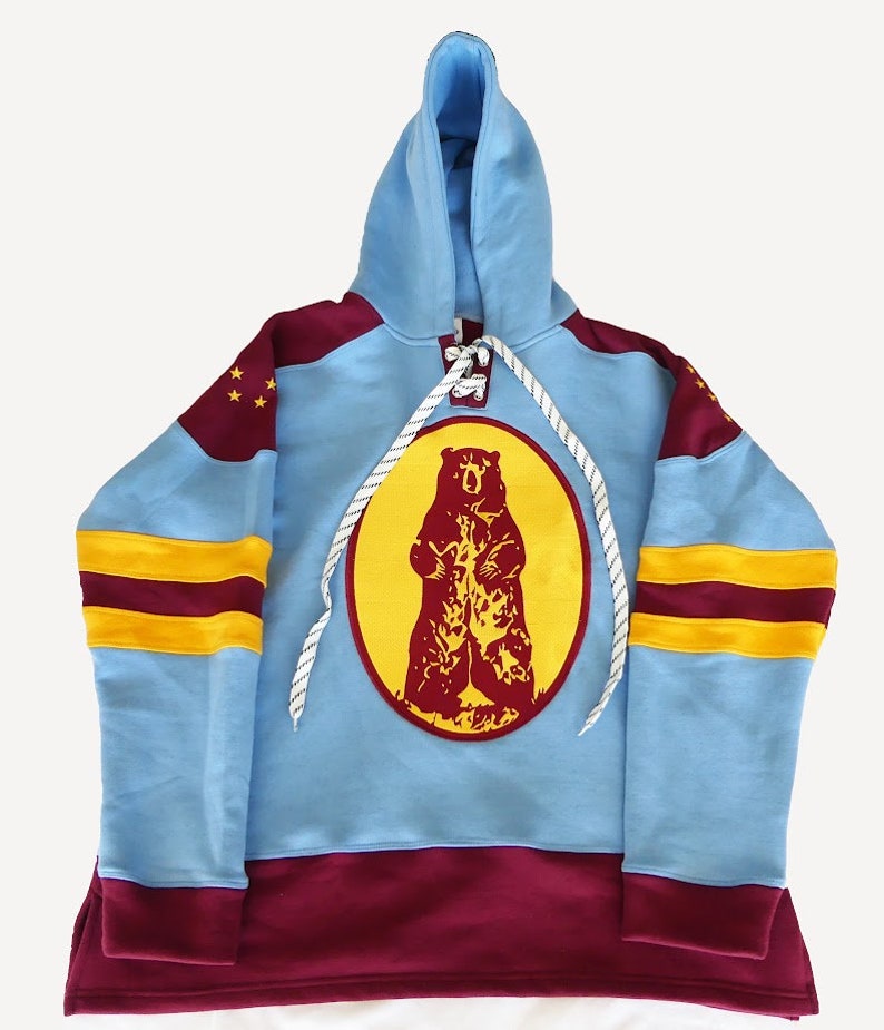 May include: A light blue hoodie with a maroon and gold hockey jersey design. The hoodie features a large embroidered bear logo on the front.