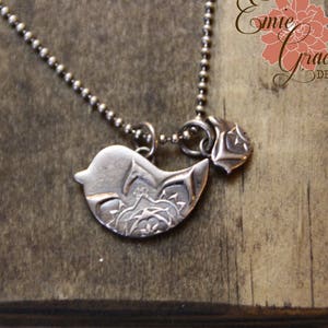 May include: A silver necklace featuring a bird pendant and a small charm. The bird pendant has an intricate design and the charm is shaped like a flower.